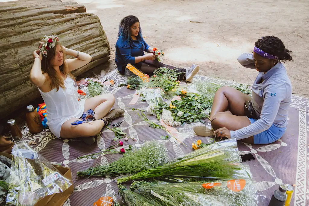 Three women sitting on a patterned rug under a fallen log, making flower crowns and arranging flowers together